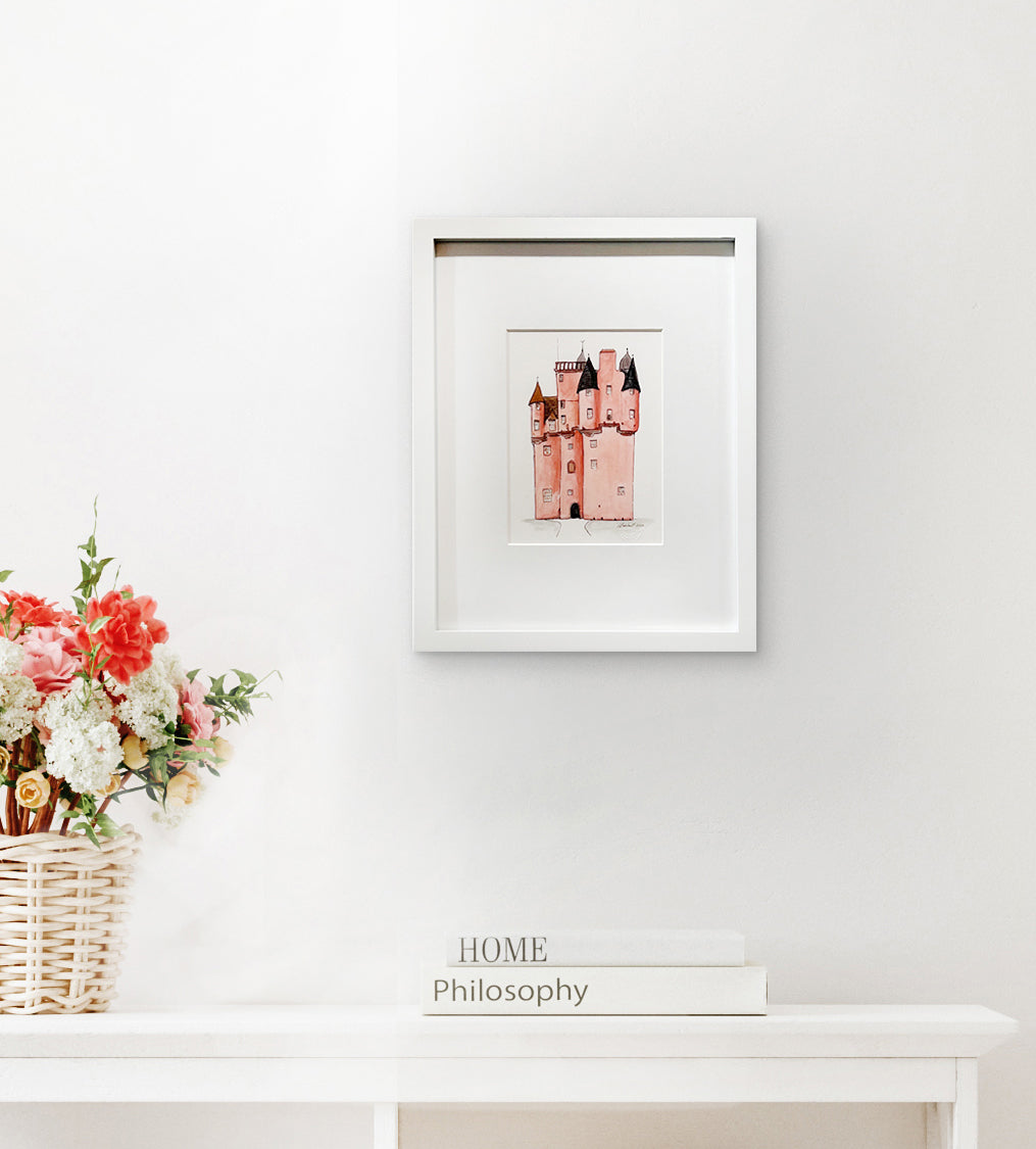 A pink, grey and brown illustration drawing of a castle in a white frame with a mat hangs on a white wall above a white surface with a stack of books and a white wicker basket with a multicolored floral arrangement.