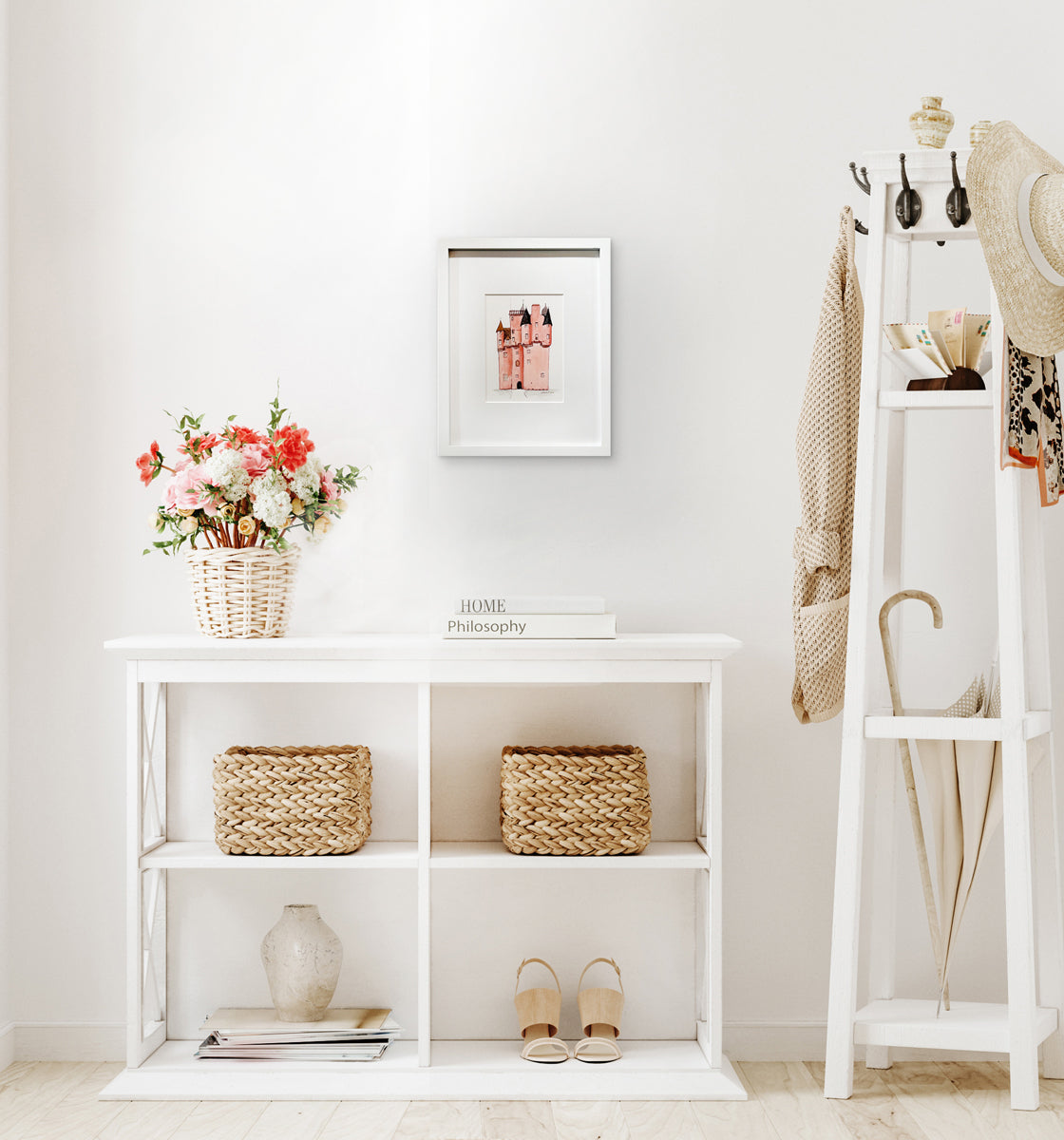 A pink, grey and brown illustration drawing of a castle in a white frame with a mat hangs on a white wall above a white shelf with a stack of books, two natural wicker baskets and a white wicker basket with a multicolored floral arrangement.