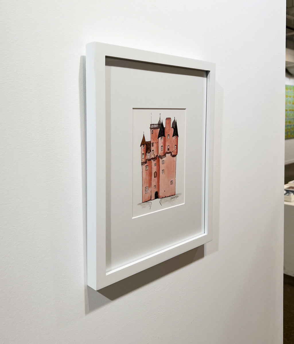 A left side-angle view of a pink, grey and brown illustration drawing of a castle in a white frame with a mat hangs on a white wall. 
