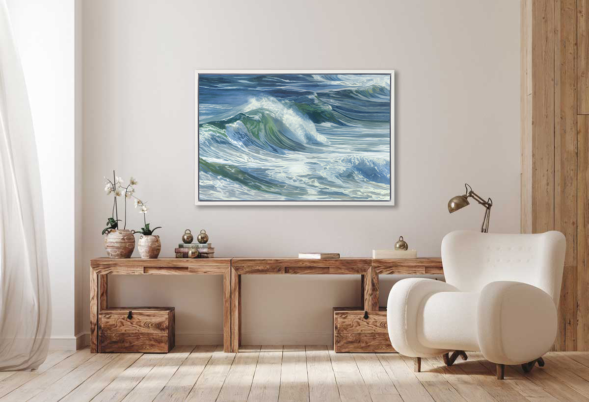 A blue, white and green abstract wave print in a white floater frame hangs on a white wall above a long wooden desk with potted orchids, stacked books and a white modern chair in front.