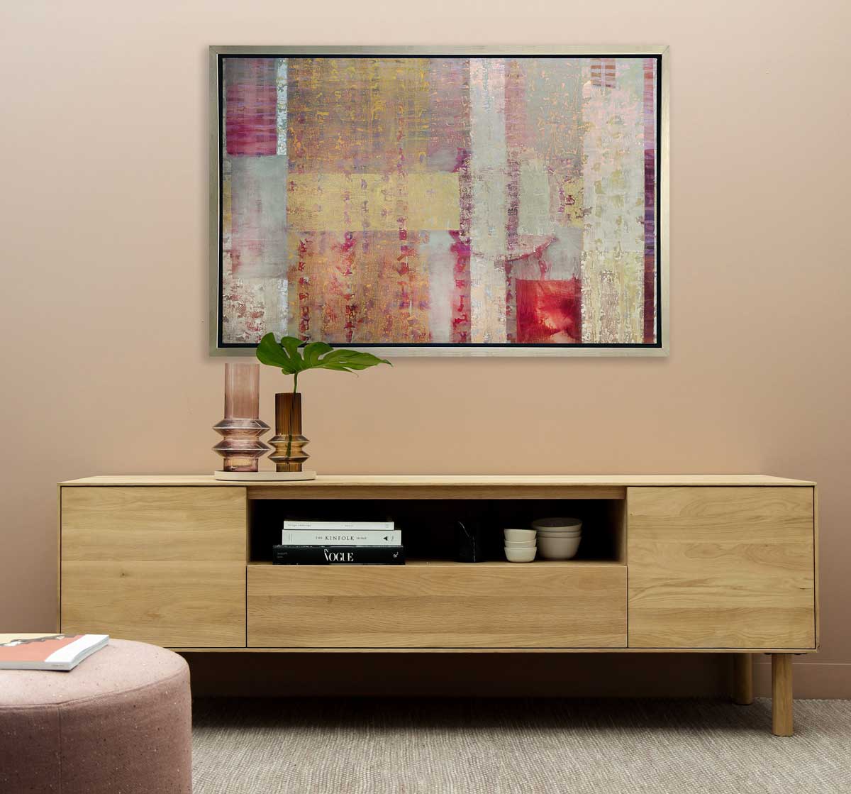 A gold, grey and red abstract print in a silver floater frame hangs on a beige wall above a wooden console table with books and vases and next to a pink ottoman.