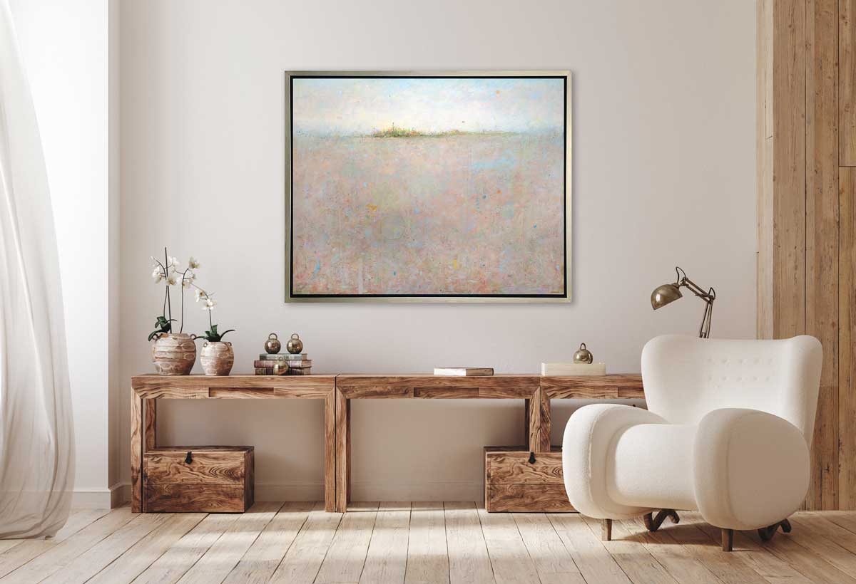 A light peach and light blue abstract landscape print in a silver floater frame hangs on a white wall above a long wooden desk with books, silver paperweights, a desk lamp, two potted white orchids and a white modern chair.