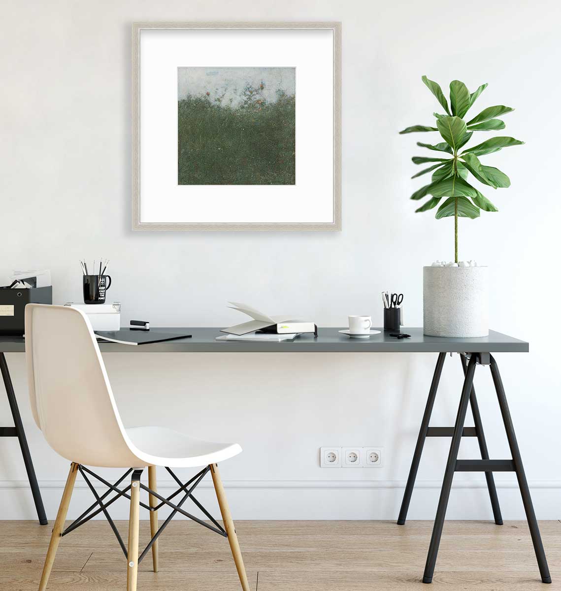 A dark green abstract landscape print in a silver frame with a mat hangs on a white wall above a dark grey desk with open notebooks, a black coffee mug with pencils and a potted plant.