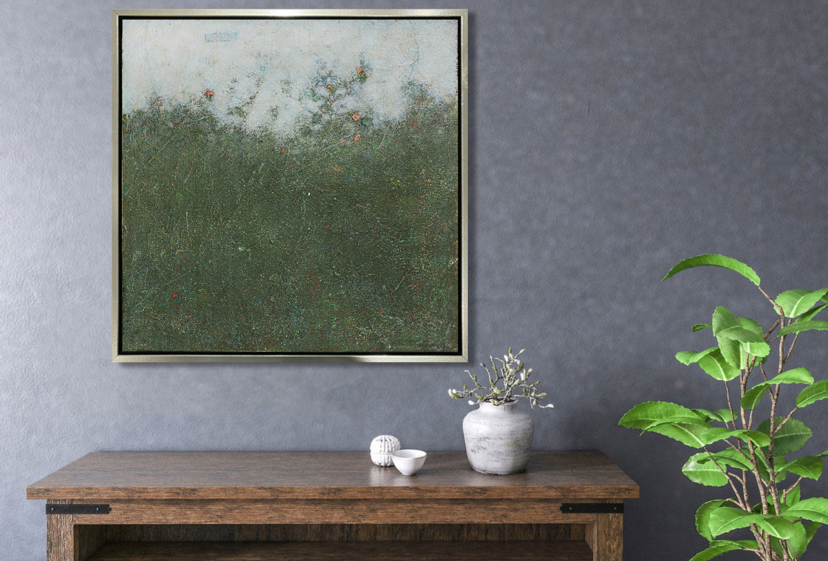 A dark green abstract landscape print in a silver floater frame hangs on a textured grey wall above a wooden cabinet with two small decorative bowls and a short vase with floral stems. 