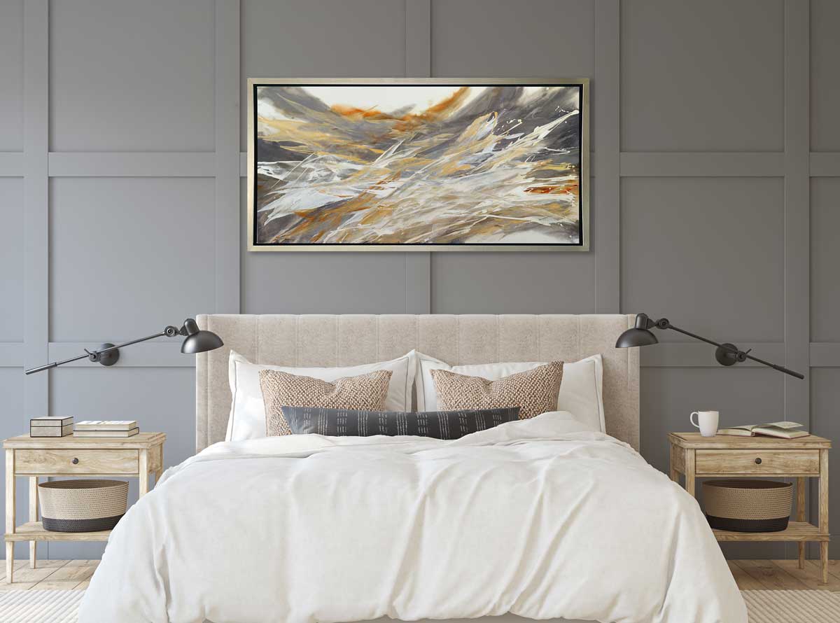 A gold, beige, white and grey abstract print in a silver floater frame hangs on a grey wall above a bed with a beige headboard, white bedding with neutral pillows and matching natural wood side tables with stacks of books.