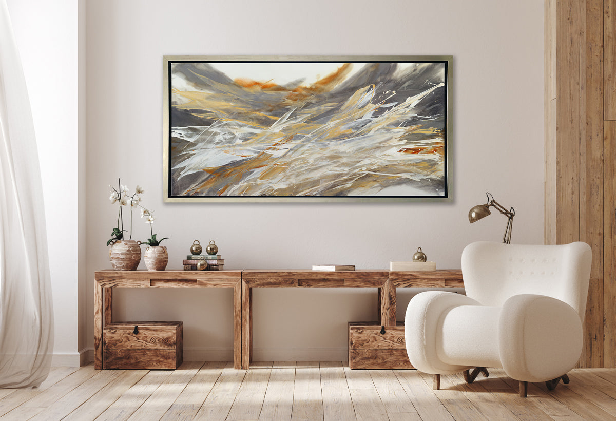 A gold, beige, white and grey abstract print in a silver floater frame hangs on a white wall above a long wood desk with two potted white orchids, decorative accessories, a chrome desk lamp and a white modern chair.