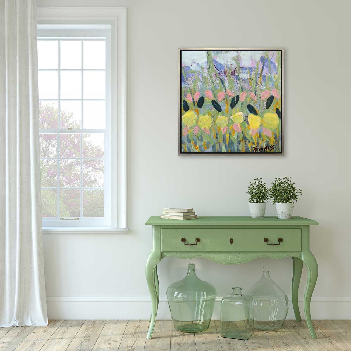 A green, yellow and pink abstract floral print in a silver floater frame hangs on a white wall above a light green console table with a stack of books, two potted shrubs and three light green glass bottles.