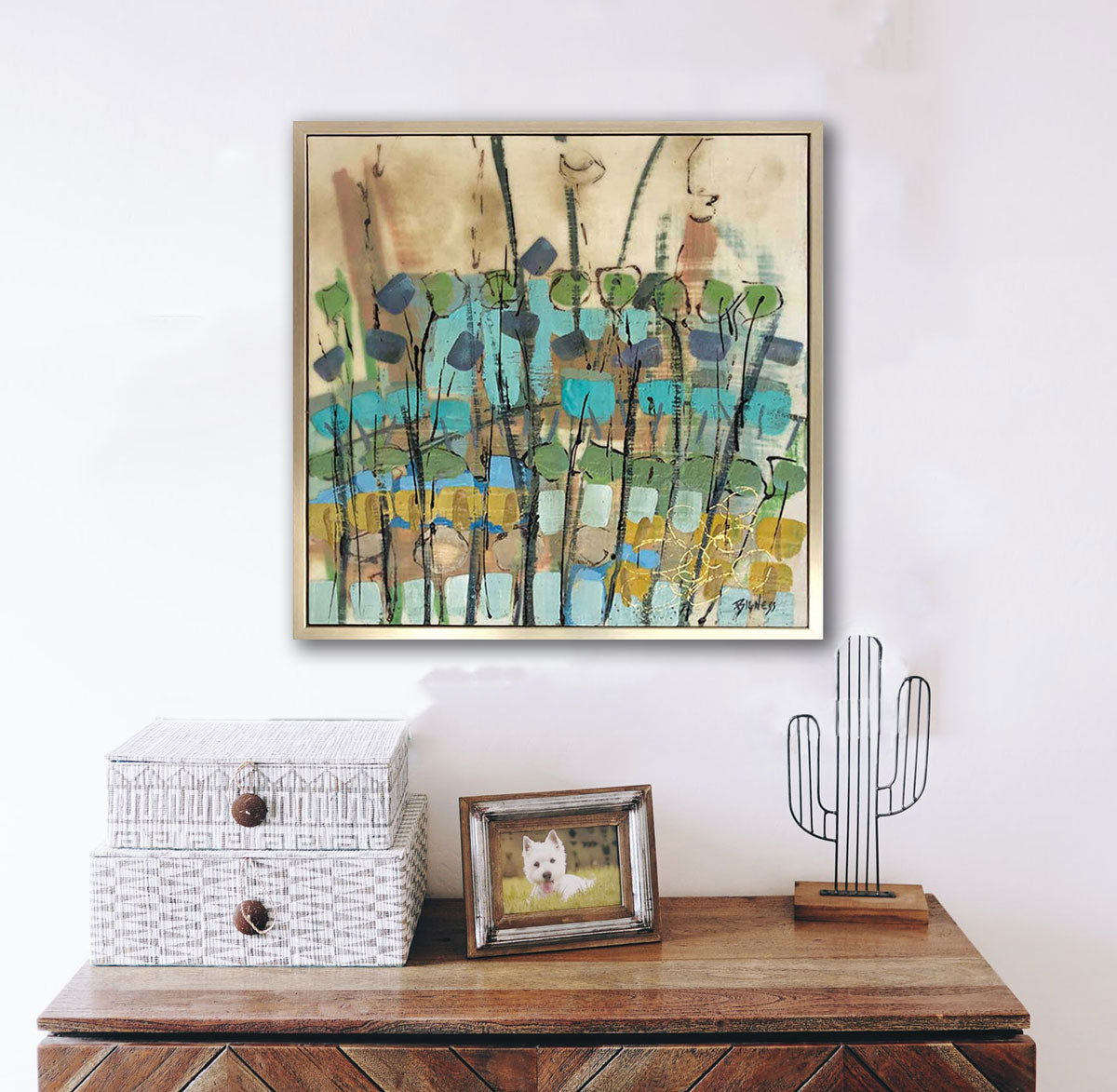 A natured-inspired abstract encaustic painting, framed in a gold floater frame, hangs on a white wall above a wooden console table with accessory boxes, a framed photo of a dog, and a cactus sculpture. 