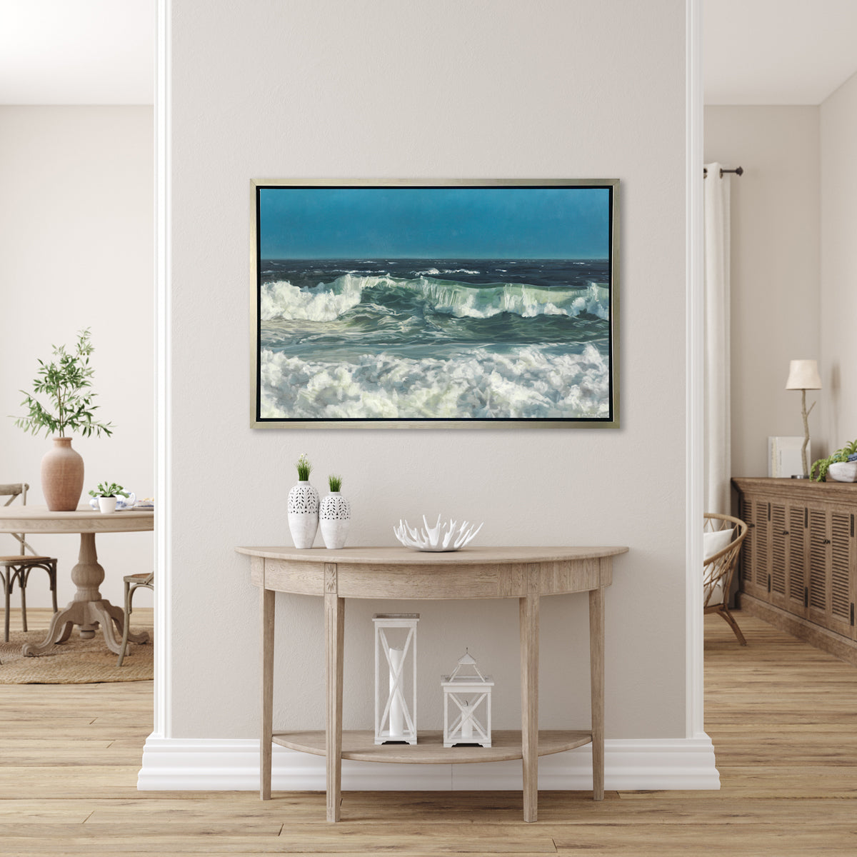 A blue, green and white abstract ocean waves print in a silver floater frame hangs on a white wall above a natural wood half moon console table with a decorative white bowl, two small white vases, and two white lanterns.