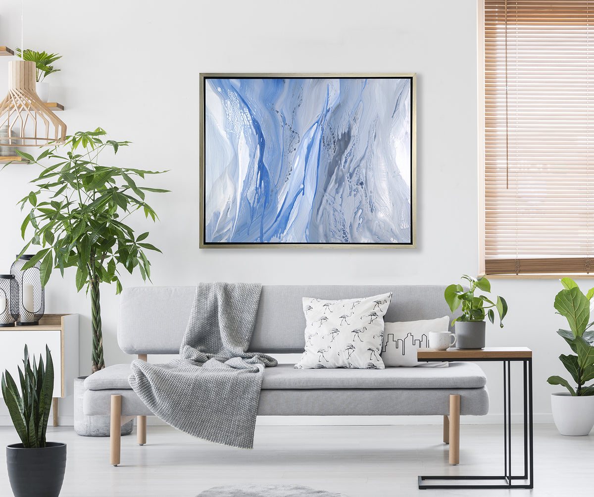 A blue, white and grey abstract print in a silver floater frame hangs on a white wall above a grey sofa with pillows, a wood side table with a potted plant and three potted floor plants.