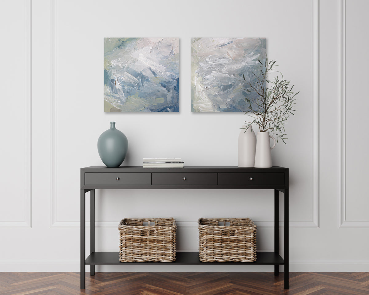 A pair of paintings are hanging on a wall over a dark brown mid century modern table with a sea-foam green vase and books on it. Its surface is decorated with white, blue and green paint in a thick impasto. Wired and ready to hang.