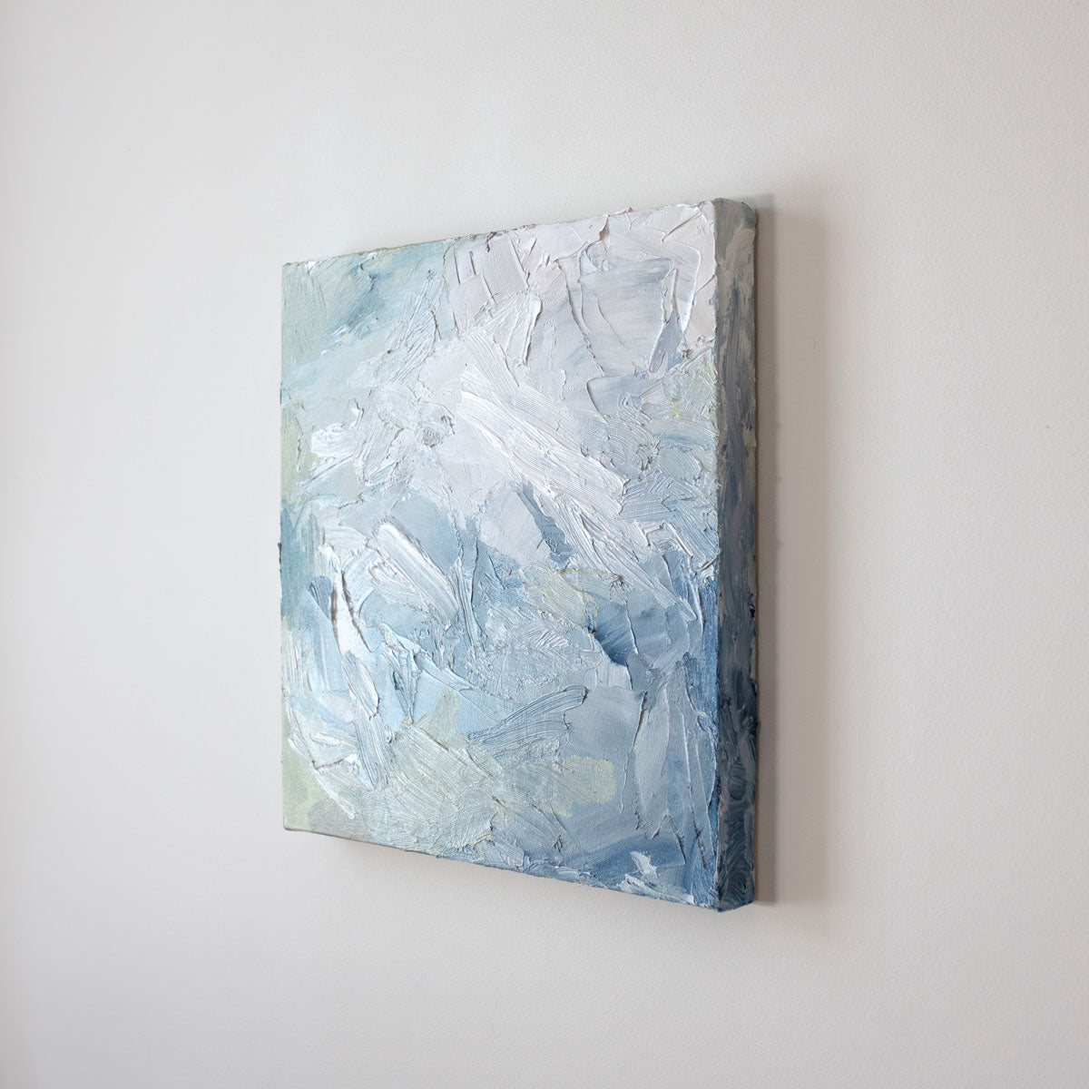 A painting is shown hanging on the wall at an angle. Its surface is decorated with white, blue and green paint in a thick impasto. Wired and ready to hang.