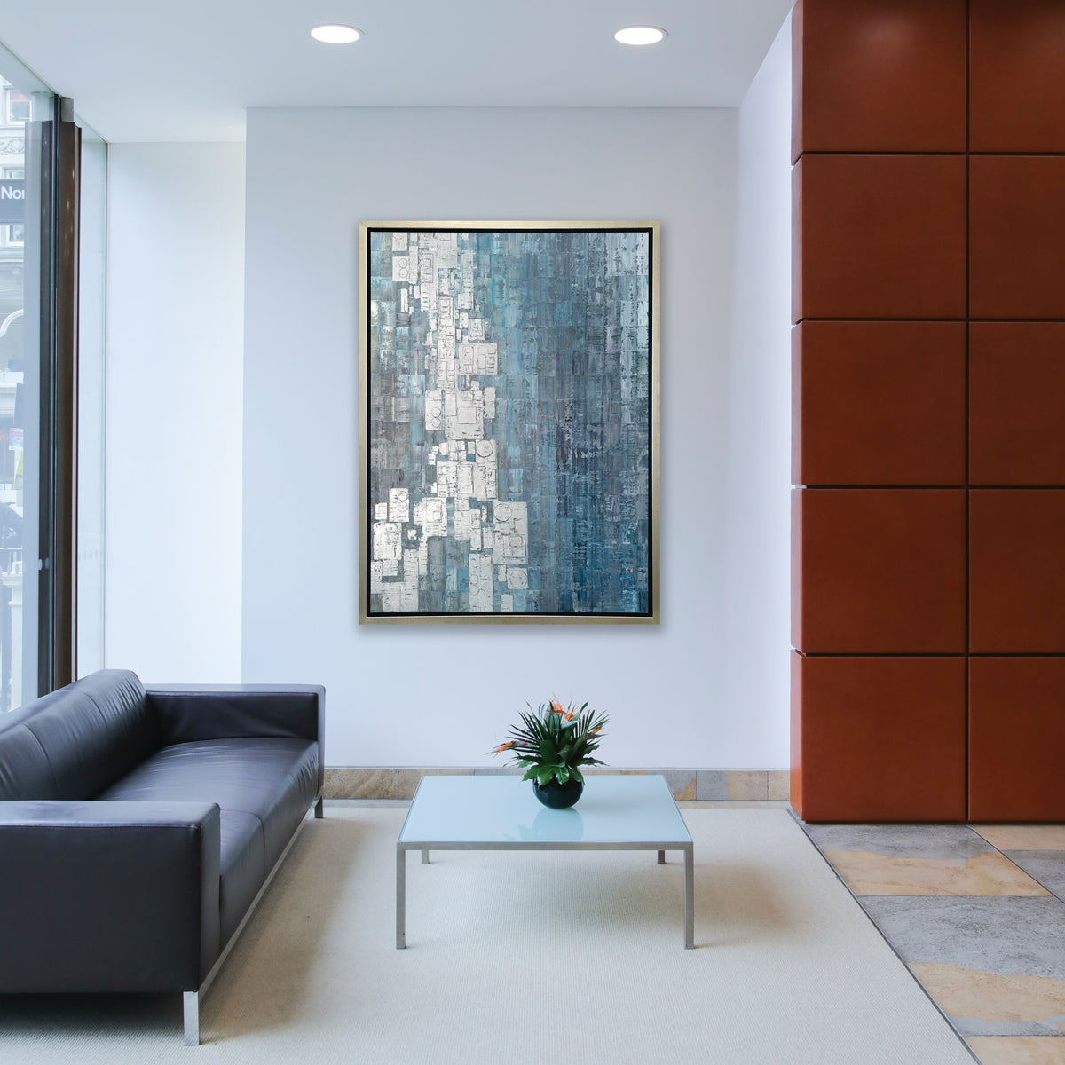 A blue, white and grey abstract print in a silver floater frame hangs on a white wall across from a dark grey sofa and a square frosted glass coffee table with a potted floral arrangement.