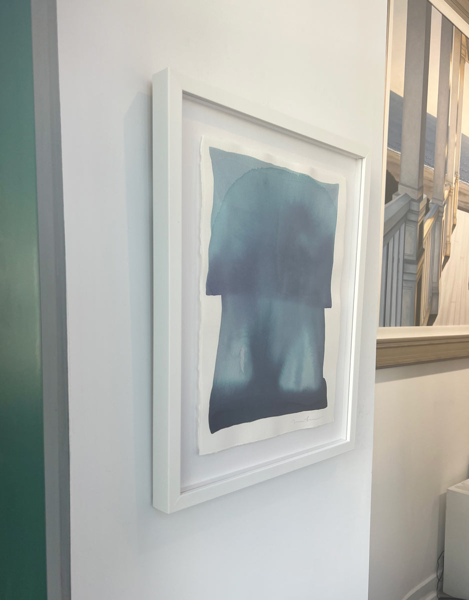 A left side-angle view of a tonal blue painting with abstract shapes by Nealy Hauschildt in a white frame floating on a white matte on a white wall.