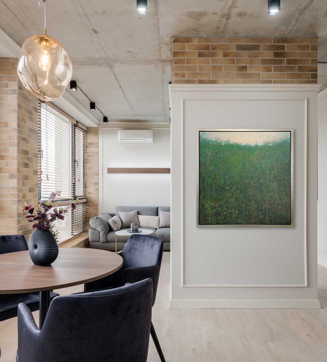A green and blue abstract landscape print in a silver floater frame hangs on a white paneled wall next to a round wooden table with deep purple chairs and a grey sofa with pillows.