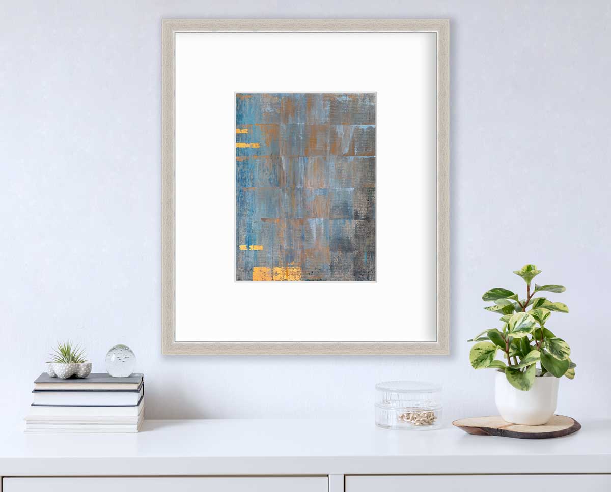 A blue, grey and gold abstract geometric print in a silver frame with a mat hangs on a white wall above a white surface with a stack of books, a clear round container with gold accessories and a potted plant.