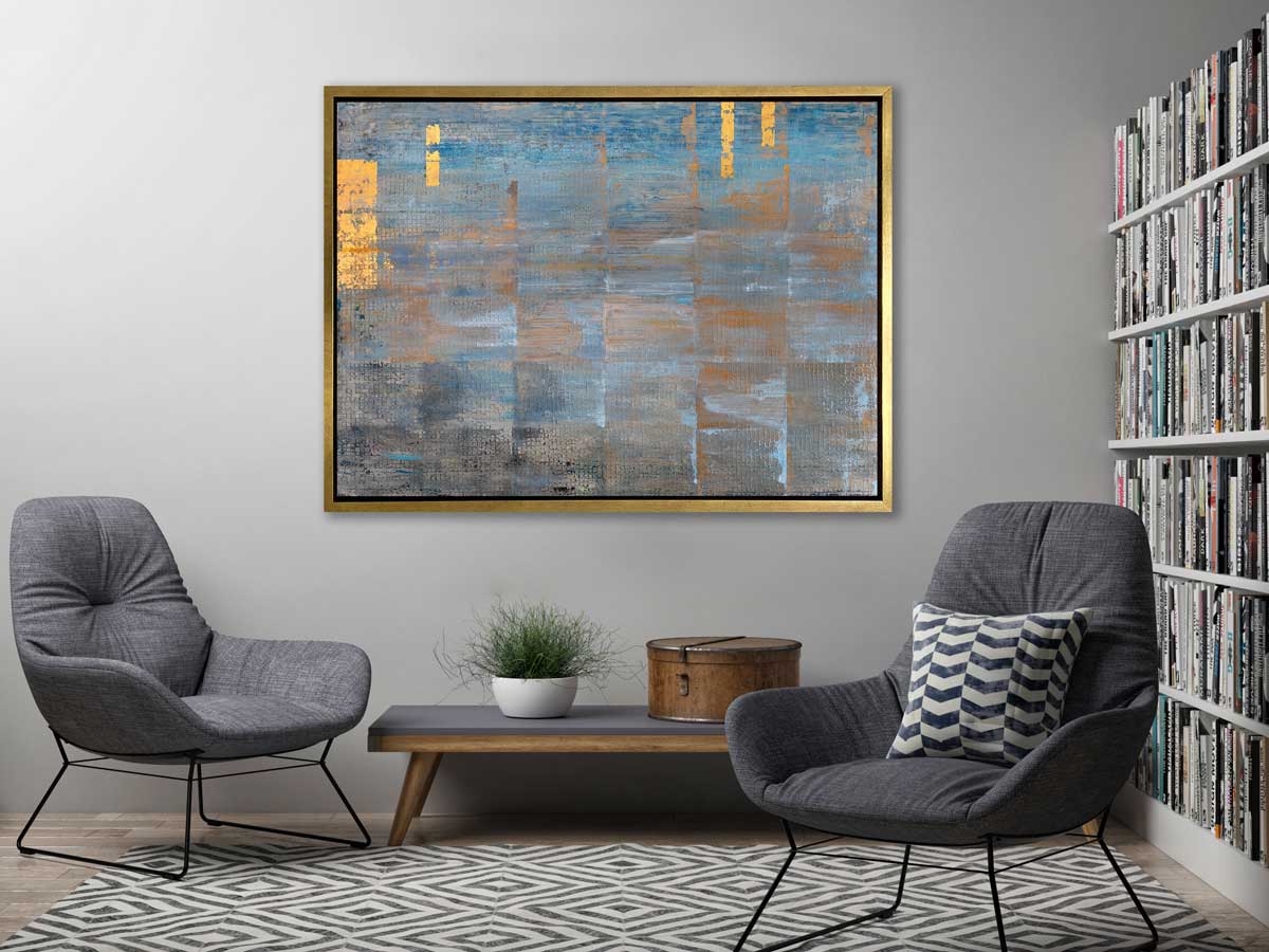 A blue, grey and gold abstract geometric print in a gold floater frame hangs on a light grey wall above a grey and wood bench with a potted plant and hat box, two grey chairs and a full wall bookshelf. 