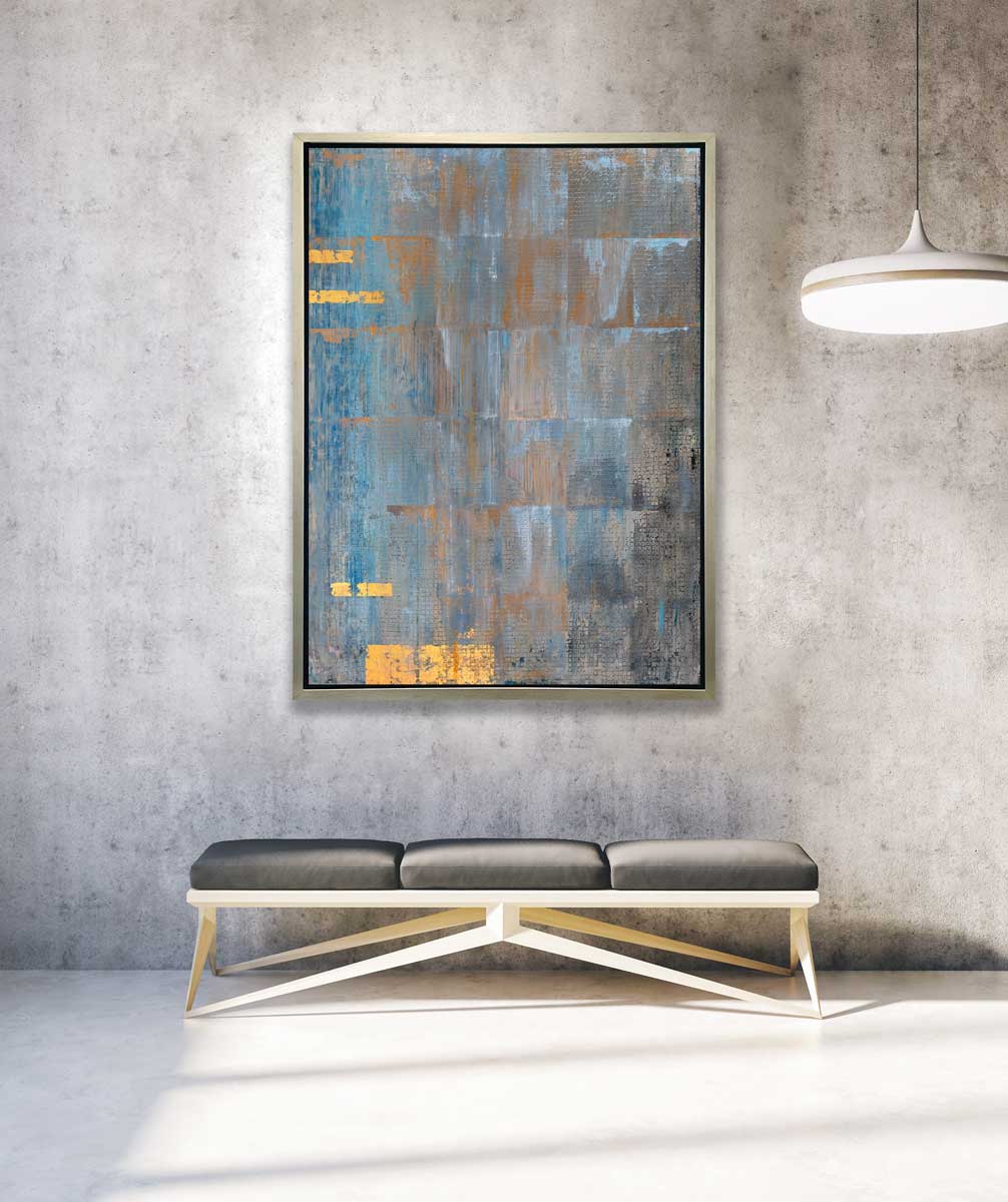 A blue, grey and gold abstract geometric print in a silver floater frame hangs on a grey industrial wall above a grey three cushioned bench.