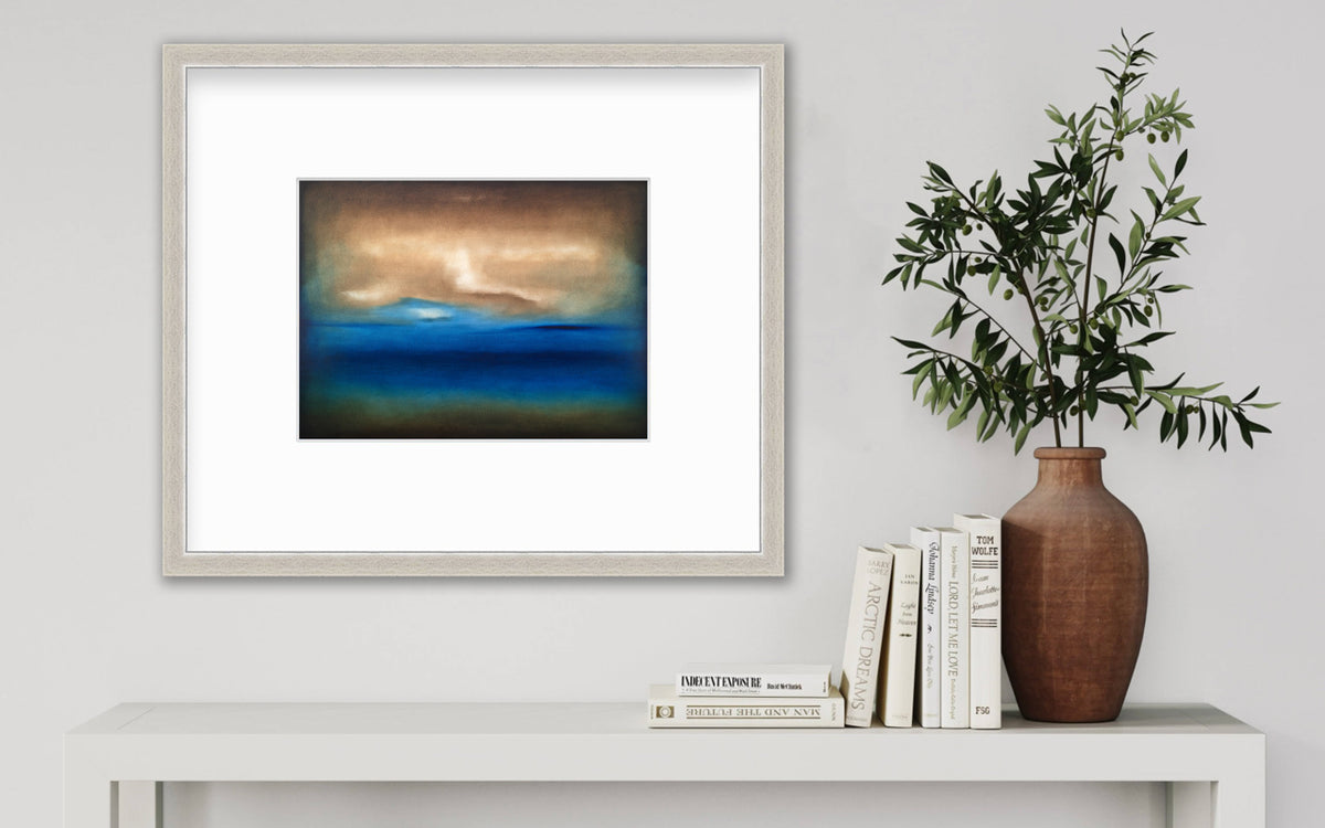 A dark brown, blue, white and teal abstract landscape print in a silver frame with a mat hangs on a white wall above a small white console table with a stack of books and a terracotta vase filled with greenery.