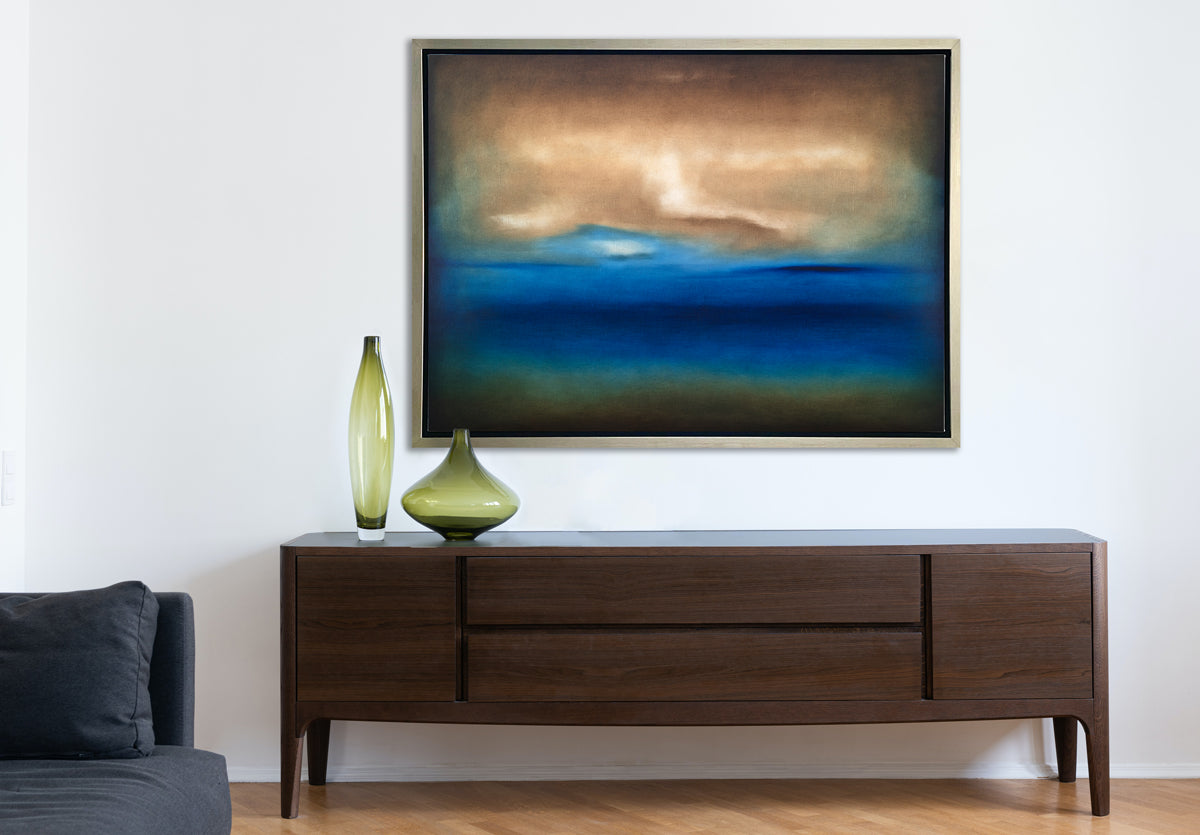 A dark brown, blue, white and teal abstract landscape print in a silver floater frame hangs on a white wall above a low dark wood credenza with two different sized and shaped green glass vases and next to a denim blue sofa.