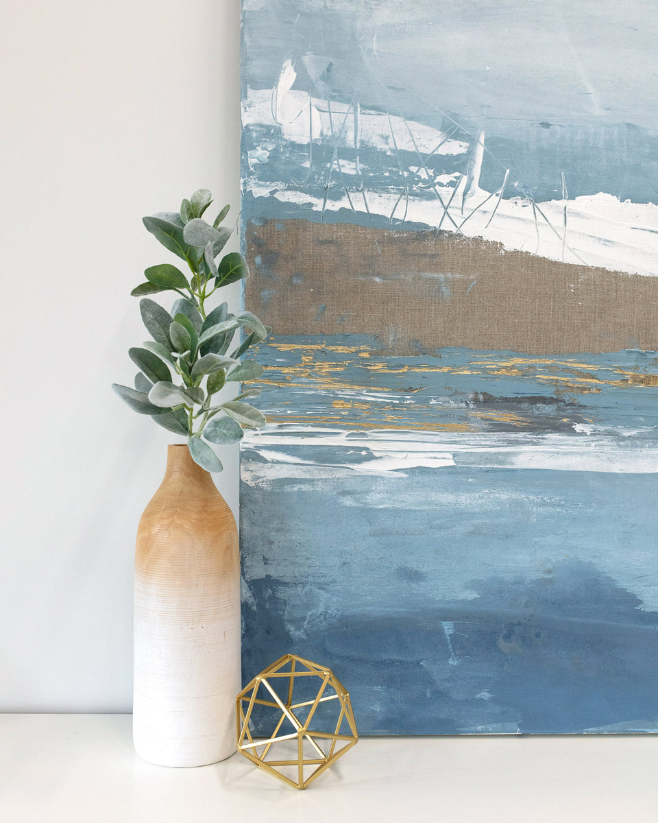 The corner of a blue abstract painting by Julia Contacessi resting on a white dresser next to modern table decor. 