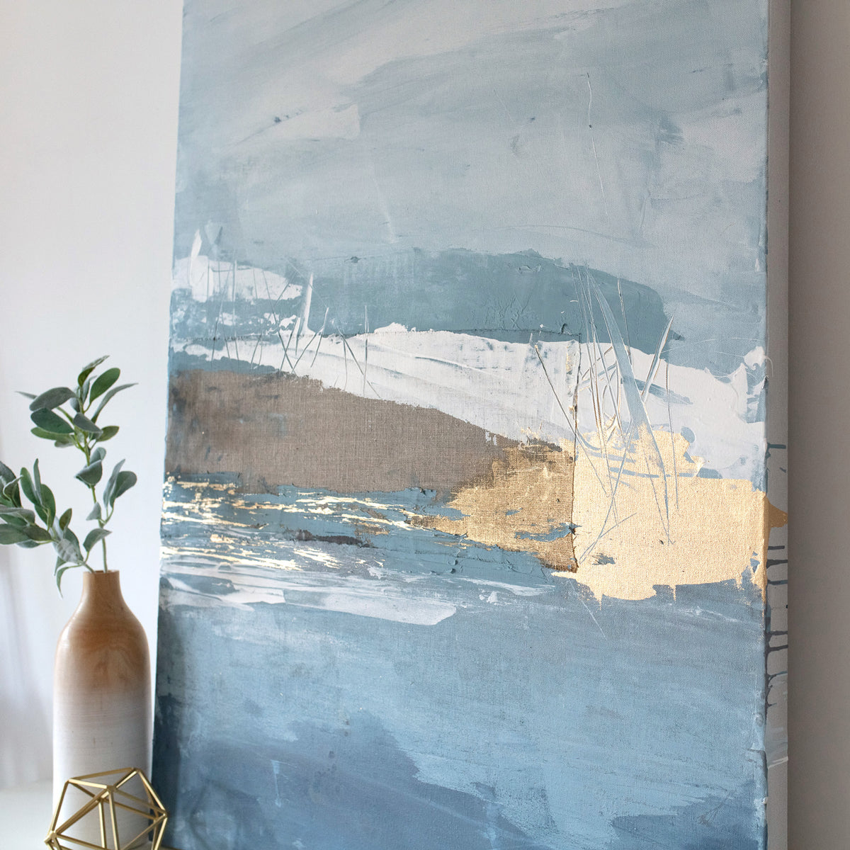 A side angle view of a blue and gold abstract painting resting on a dresser next to modern table decor. 