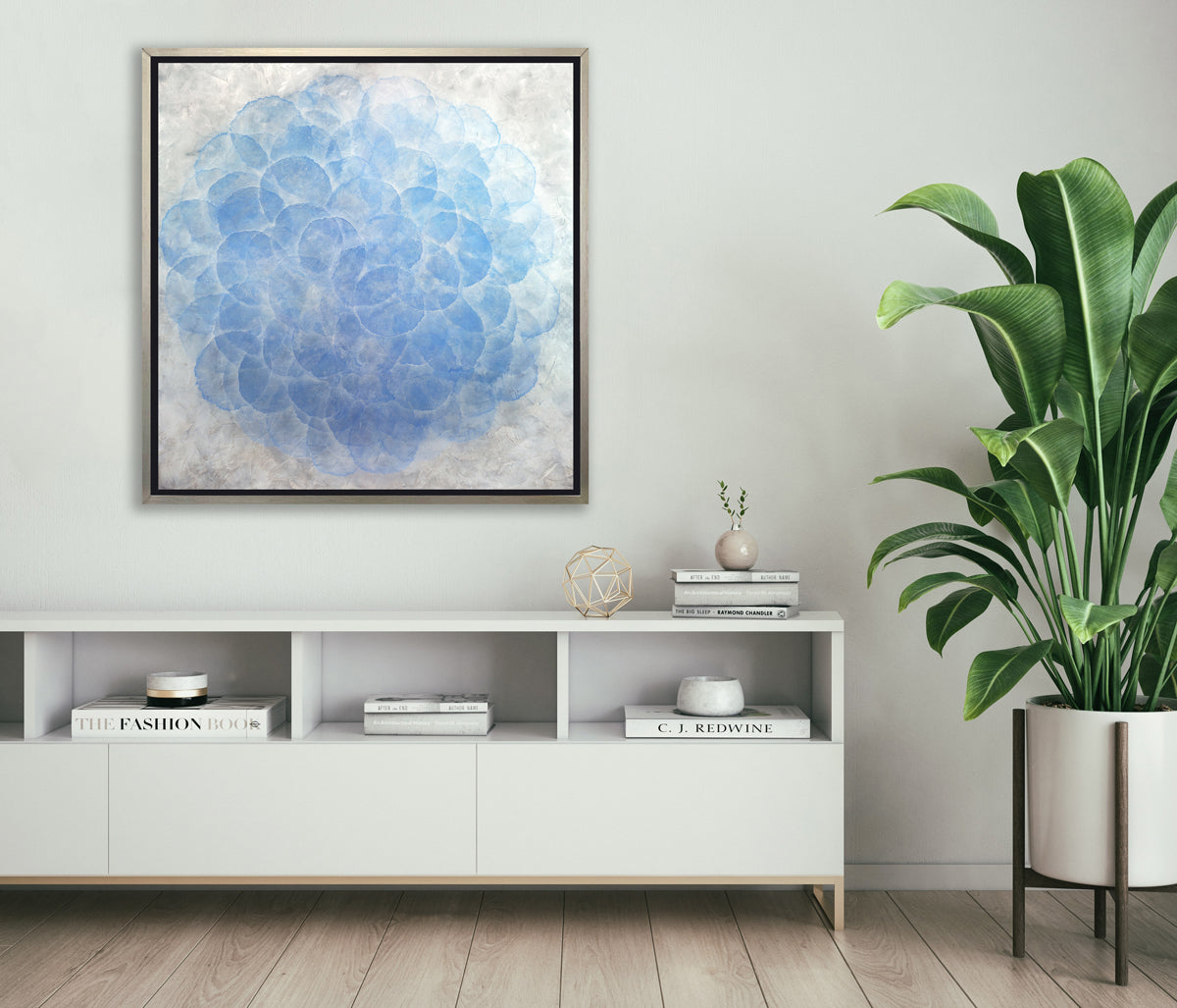 A blue, white and silver abstract geometric print in a silver floater frame hangs on a white wall above a white console shelf with stacks of books, a gold wire sphere and a large potted floor plant.