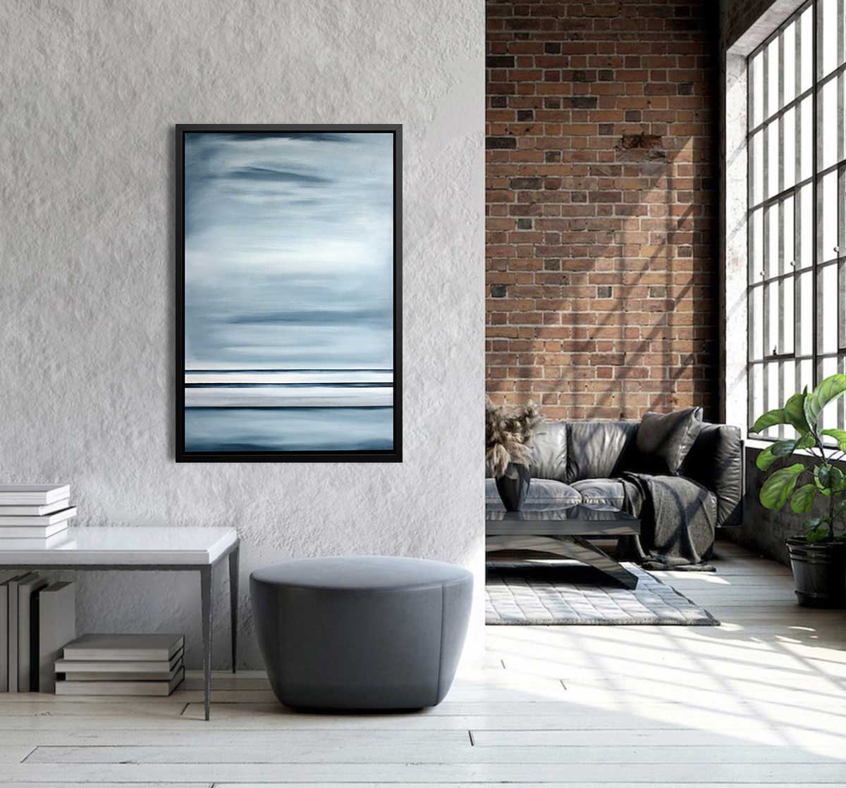 Blue-grey geometric abstract print hangs in a black floater frame on a wall above a table with books and an ottoman overlooking a living room with a brown leather couch and coffee table.