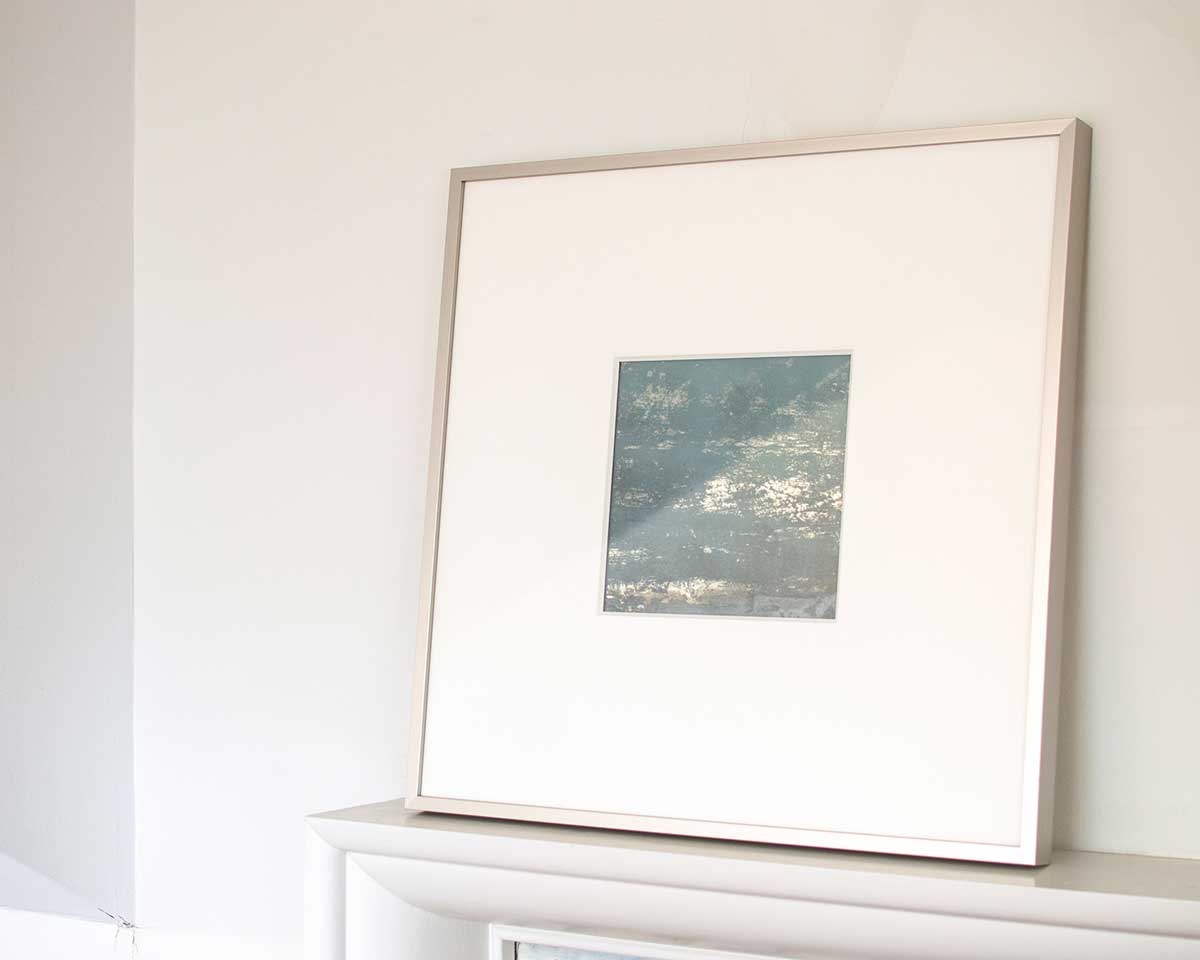 A right side-angle view of a small light blue and silver painted art mirror in a silver frame with a large white mat sits on a white surface.