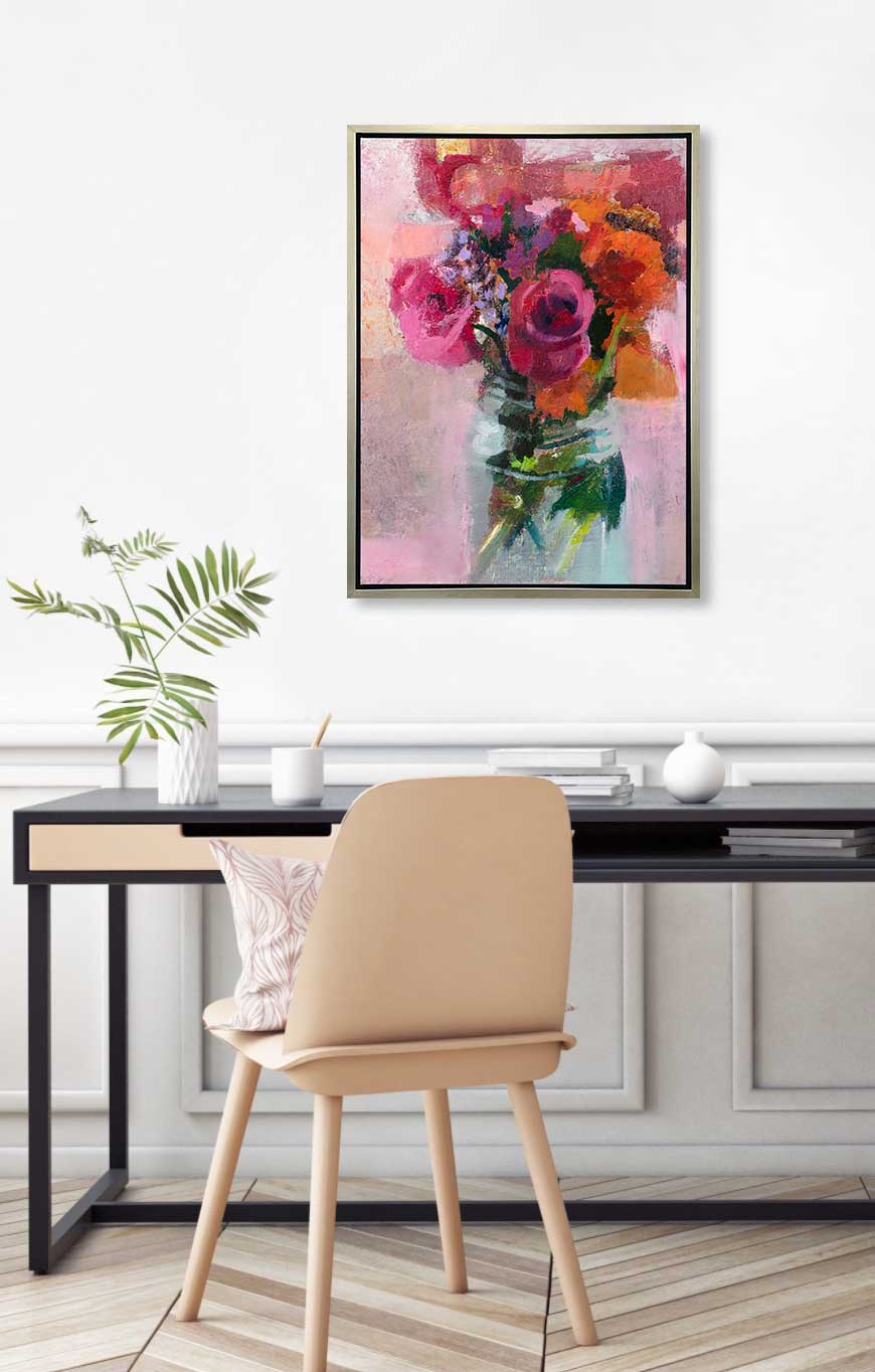 A pink, orange and green abstract floral print in a silver floater frame hangs on a white wall above a black desk with a stack of books, a white vase with a green stems and a beige chair with a pink and white printed pillow.