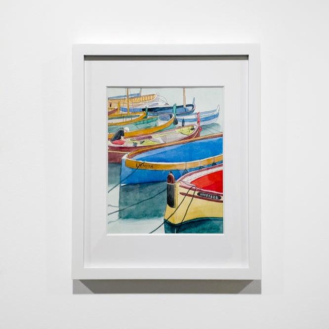 A colorful framed watercolor painting of boats docked in a marina hangs on a white wall. 