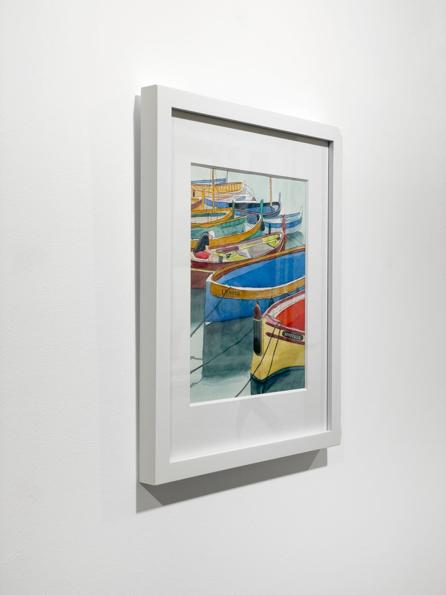 A side view of a colorful framed watercolor painting of boats docked in a marina hanging on a white wall.