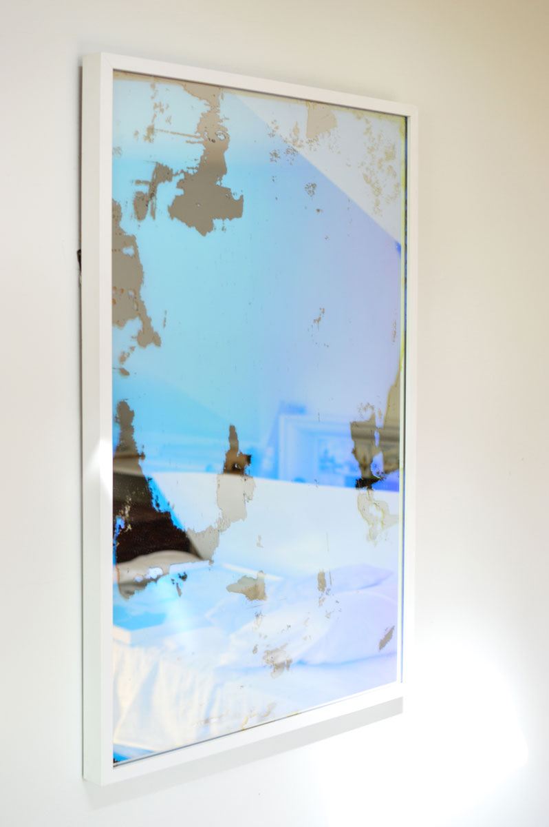 A left side-angle view of an art mirror with dichroic film by Alina B hangs on a white wall.