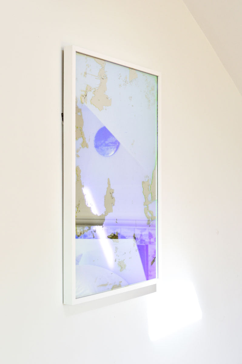 A left side-angle view of an art mirror hangs on a white wall.