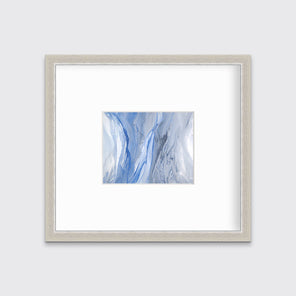 A blue, white and grey abstract print in a silver frame with a mat hangs on a white wall.