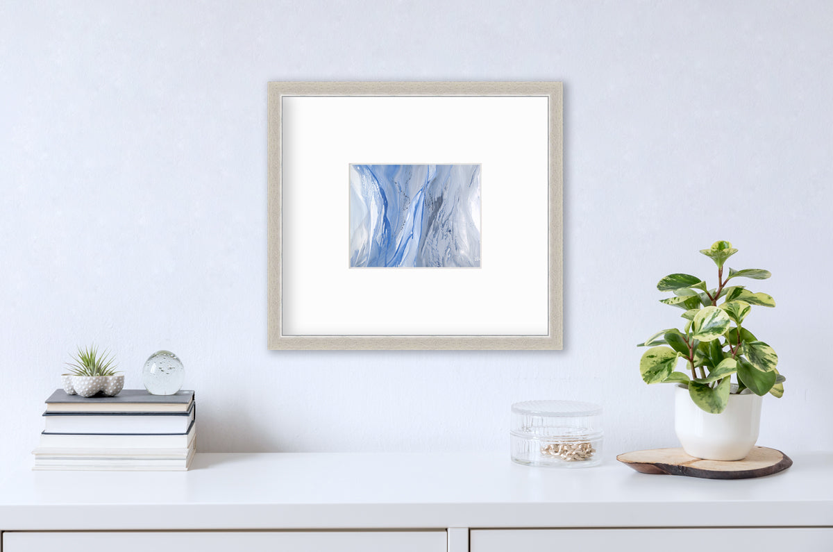 A blue, white and grey abstract print in a silver frame with a mat hangs on a white wall above a white surface with a stack of books, a round clear container with gold accessories and a potted plant.