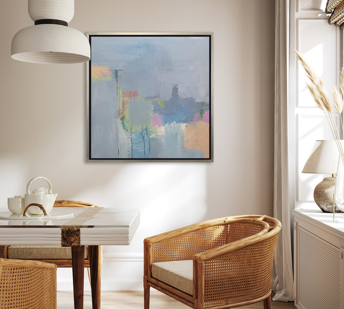 A slate blue, green, light orange and pink abstract print in a silver floater frame hangs on a white wall above a glass table, rounded rattan chairs and a window shelf with an off white table lamp.