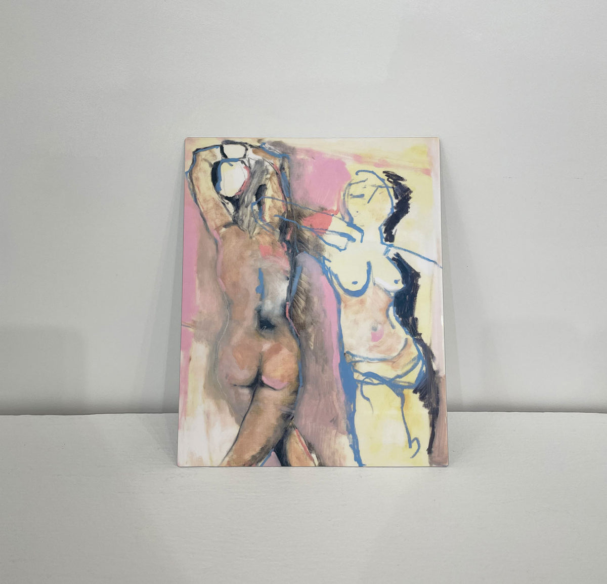 An abstract nude figurative print on metal rests propped against a white wall on a clean, white surface. 