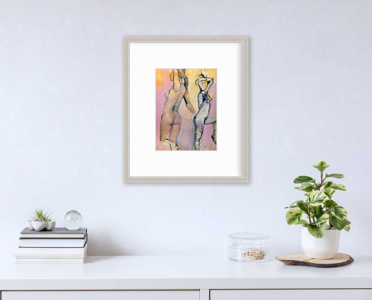 A pink, yellow, blue and black abstract figurative print of two women in a silver frame with a mat hangs on a white wall above a white surface with a stack of books, a round clear container with gold accessories and a potted plant.