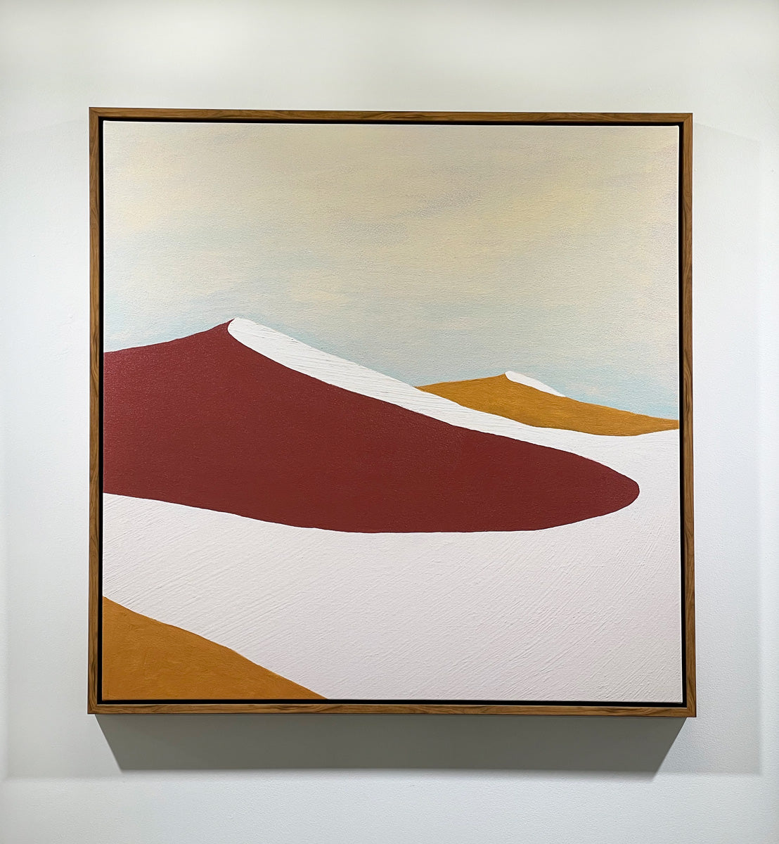 A red, burnt orange and beige abstract print in an oak wood floater frame hangs on a white wall.
