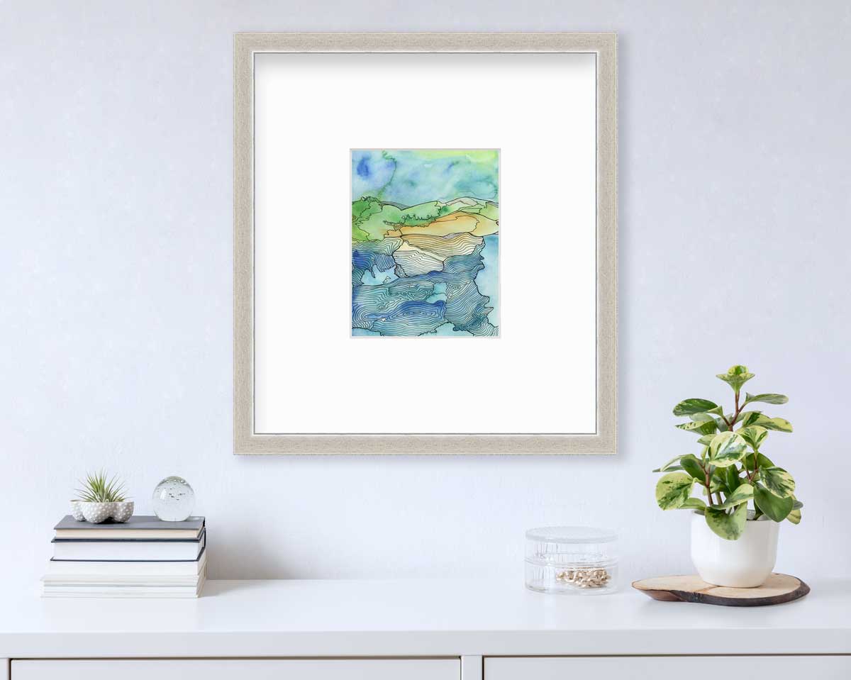 A blue and green landscape illustration in a silver frame with a mat hangs on a white wall above a white surface with a stack of books, a clear round trinket container with gold accessories and a potted plant.