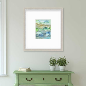 A green and blue landscape illustration print framed in a silver frame with a mat hangs on a white wall above a green console table with a stack of books and two potted plants.
