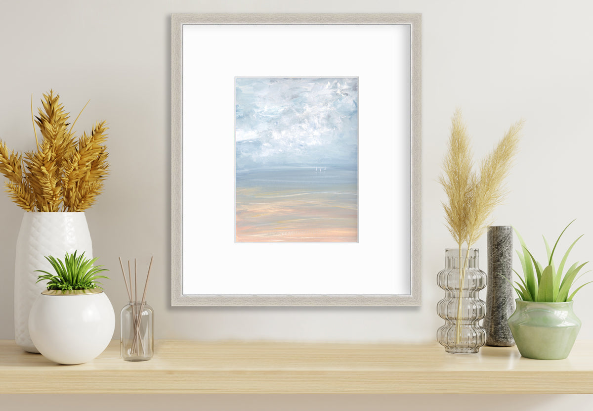 A white, blue and beige abstract seascape print in a silver frame with a mat hangs on a white wall above a wood surface with several various vases with wheat stems and greenery.