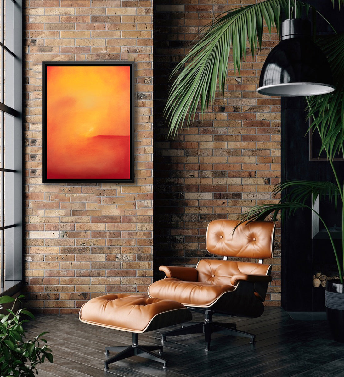 An orange and red abstract print in a black floater frame hangs on a brick wall above a tufted leather lounge chair with a matching ottoman.