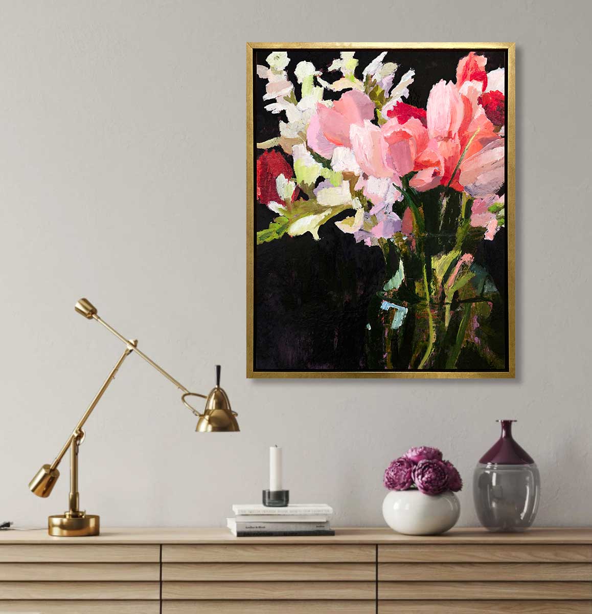 A pink, black and green abstract floral print in a gold floater frame hangs on a white wall above a wood sideboard with a gold adjustable desk lamp, a stack of books, a white round vase with purple flowers and a purple and glass vase.