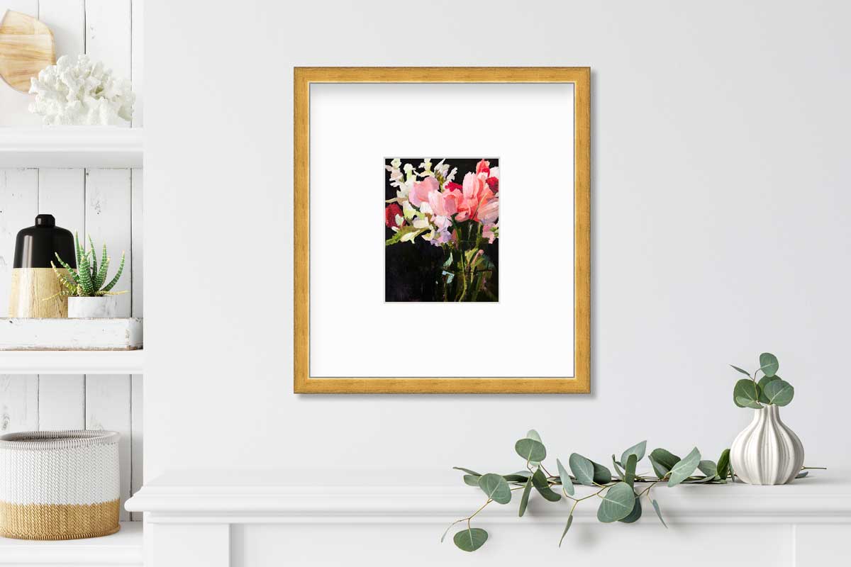 A pink, black and green abstract floral print in a gold frame with a mat hangs on a white wall above a white shelf with a small white vase and eucalyptus leaves and next to a built in books shelf with accessories.