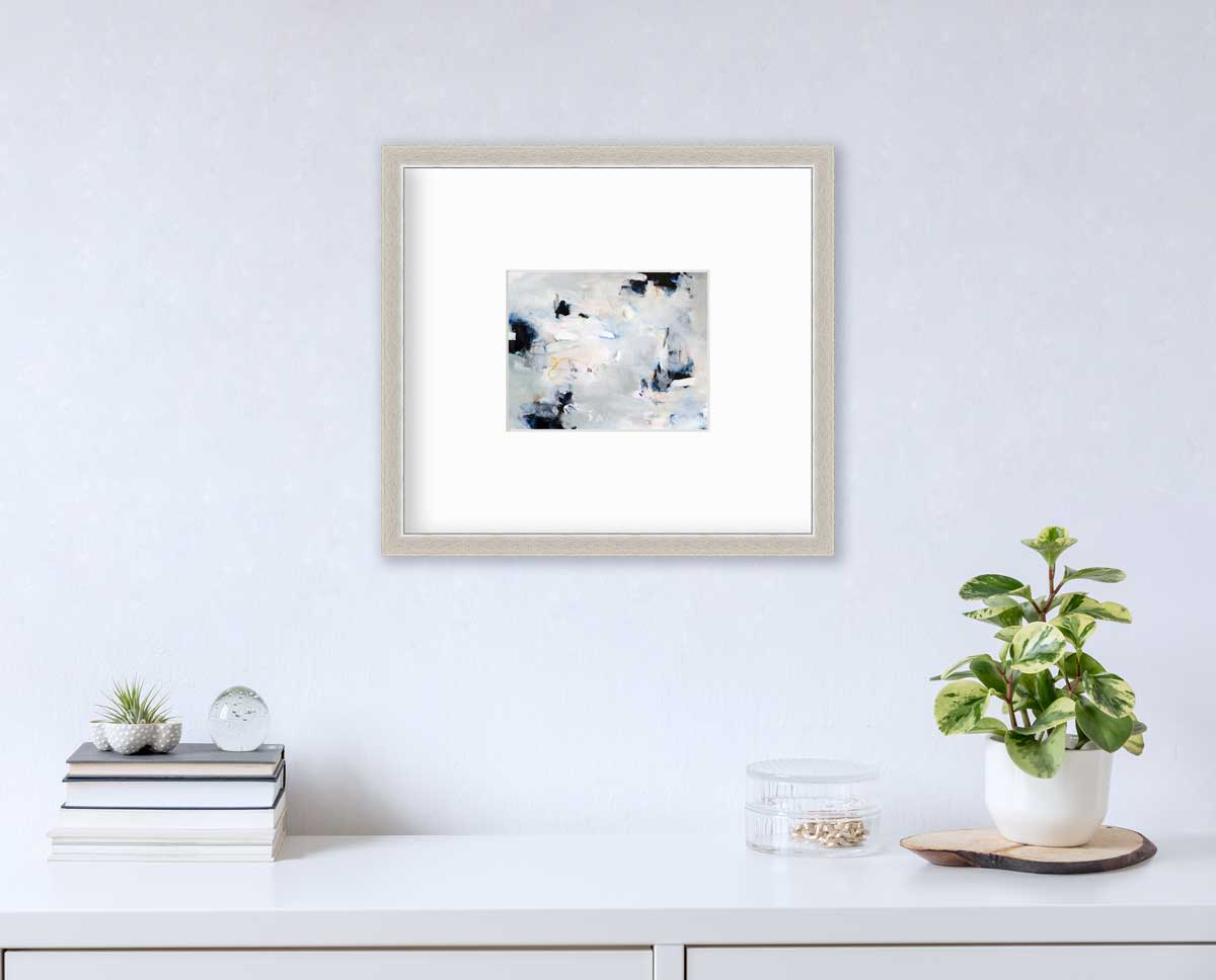 A black and white abstract print by Kelly Rossetti hangs in a silver frame with a mat on a white wall above a white surface with a stack of books, a round clear container with gold accessories and a potted plant.