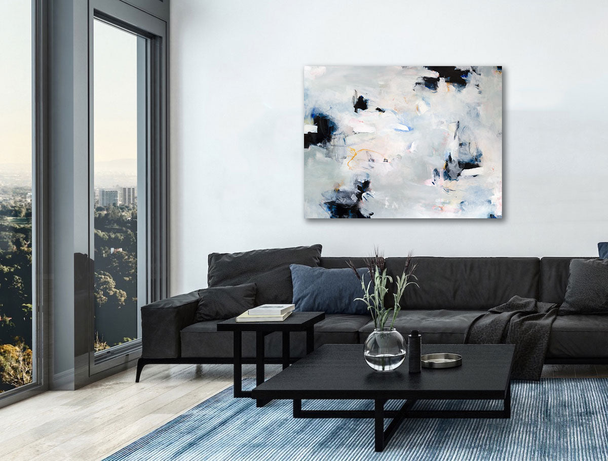A white and black abstract painting hangs above a large black sofa, a modern high-low coffee table and a blue rug. Windows to the left overlook a view of trees.