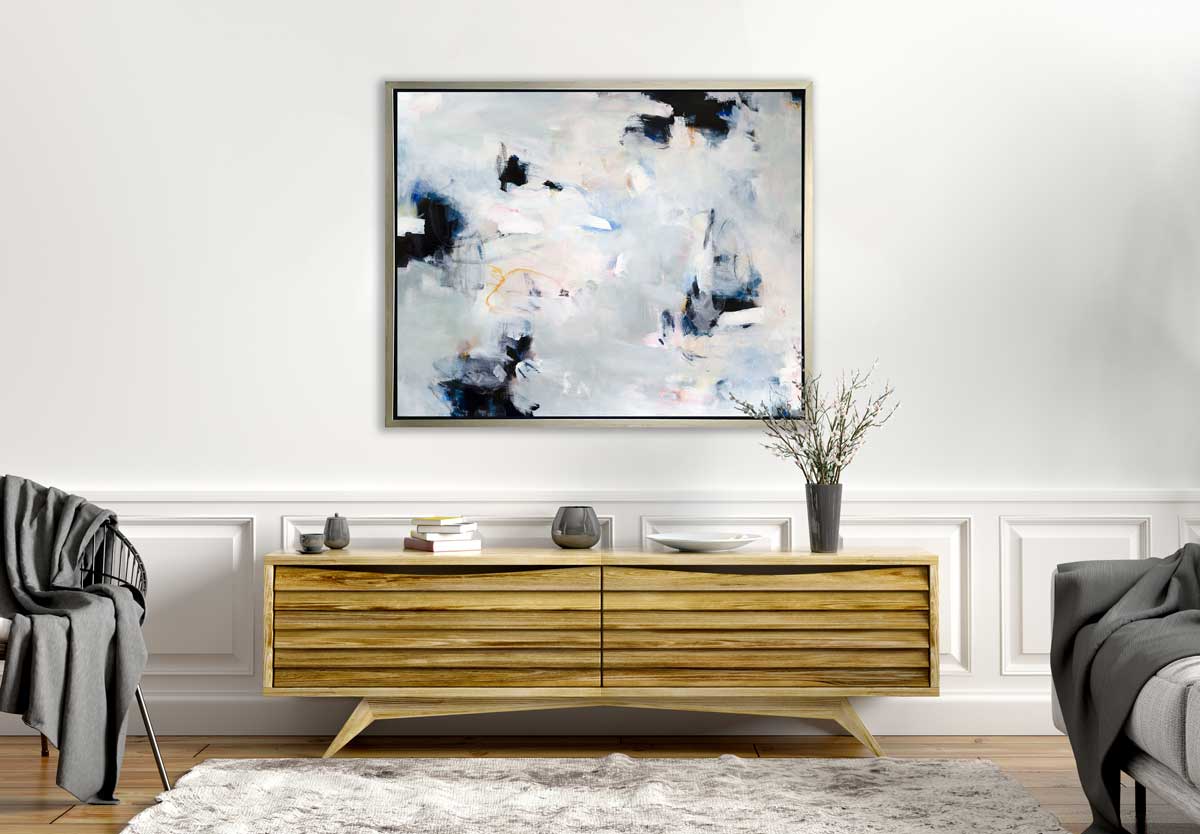 A white, black and grey abstract print in a silver floater frame hangs on a white wall above a wood cabinet with grey vases, a stack of books and a white plate.