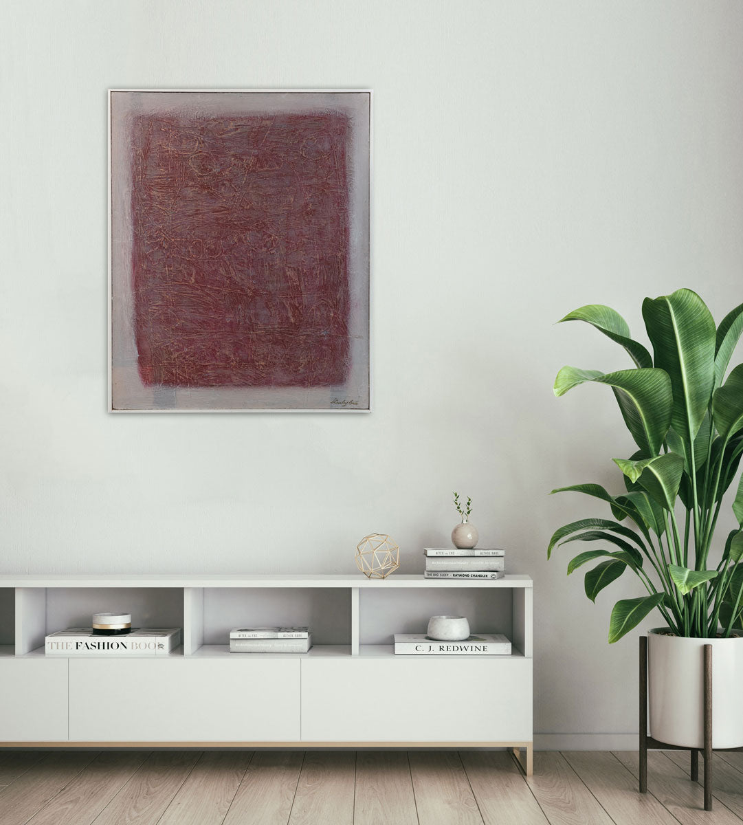 A red and white abstract painting in a white floater frame hangs on a white wall above a white credenza with books, decorative items and a floor potted plant to the right.